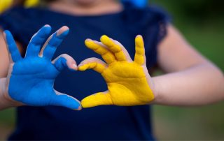 Ein Kind formt mit seinen Fingern ein Herz, während die eine Hand blau und die andere gelb bemalt ist.